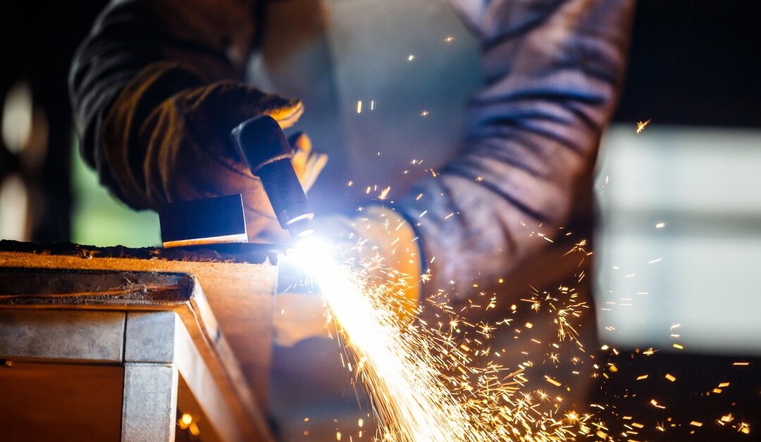 A welder uses plasma equipment to cut a thick metal sheet at their workspace, releasing a shower of hot sparks.